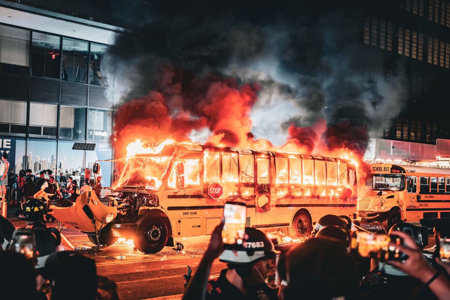 A large city bus engulfed in flames at night during a protest, surrounded by crowds of onlookers on a urban street.