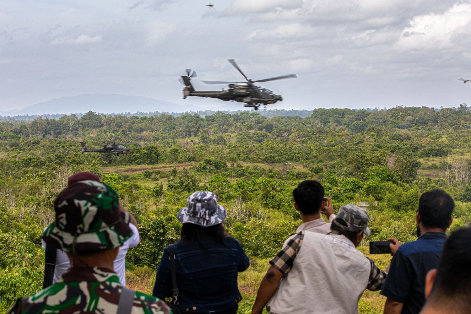 Spectators watch two military Apache helicopters fly over a lush green jungle.
