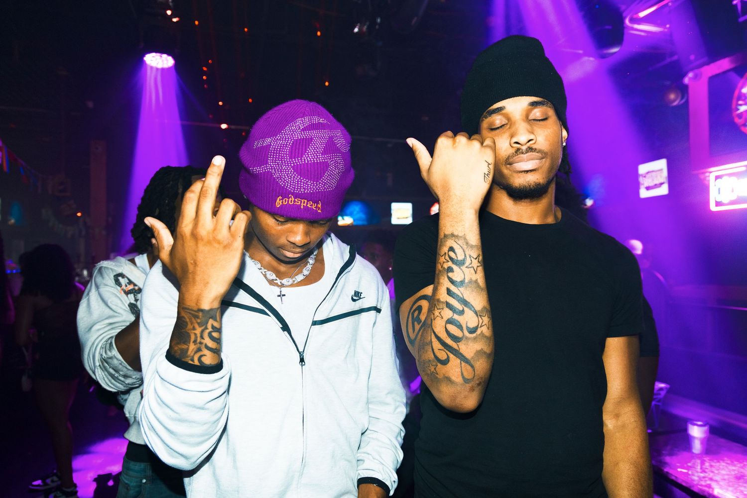 Two tattooed men posing in a nightclub with purple neon lights, one wearing a purple cap and white jacket.