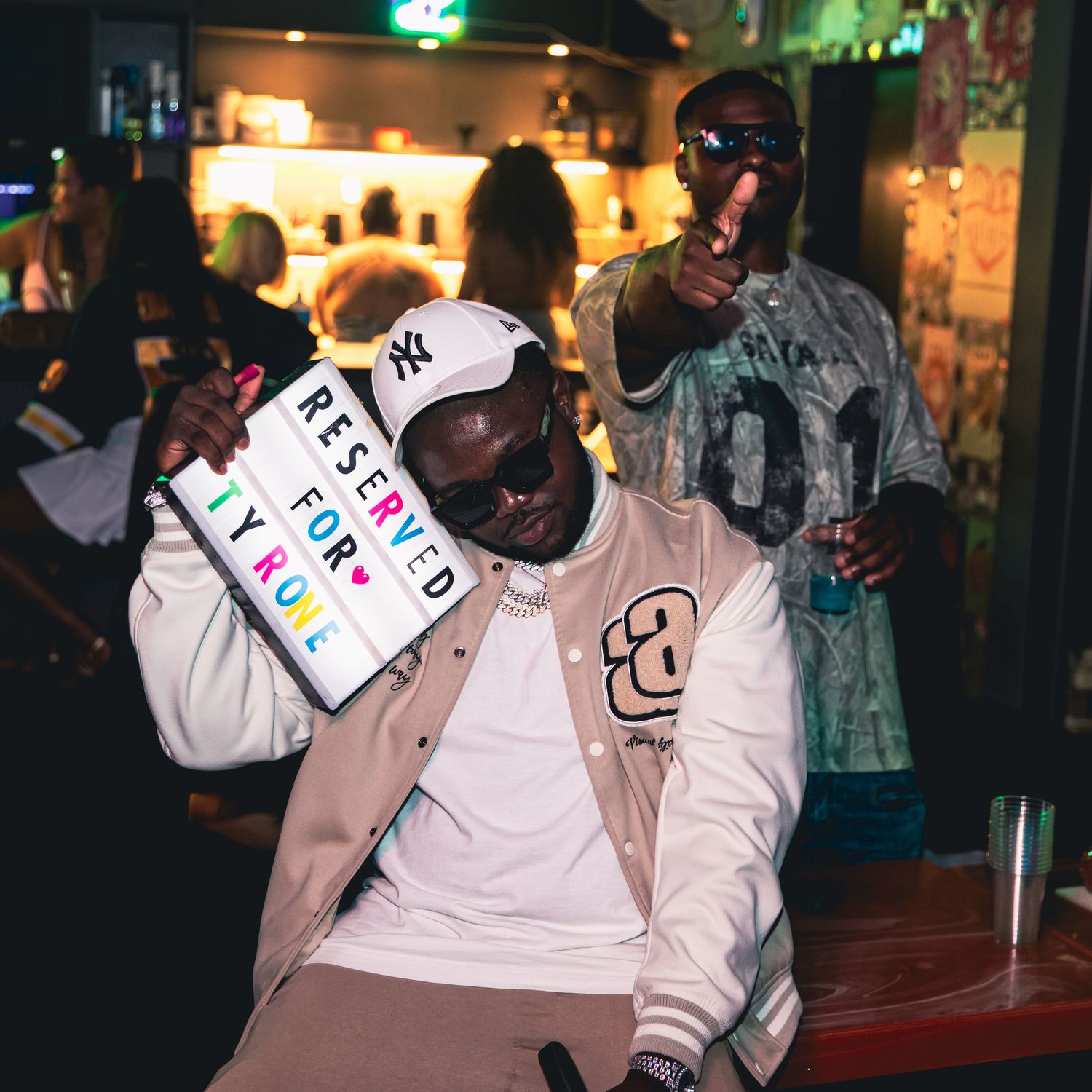 Man in NY Yankees cap holds Reserved for Tyrone sign at a lively bar party event.