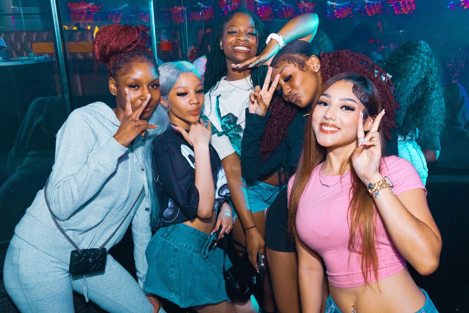 Four stylish young women pose together making peace signs at a colorful nightclub party.