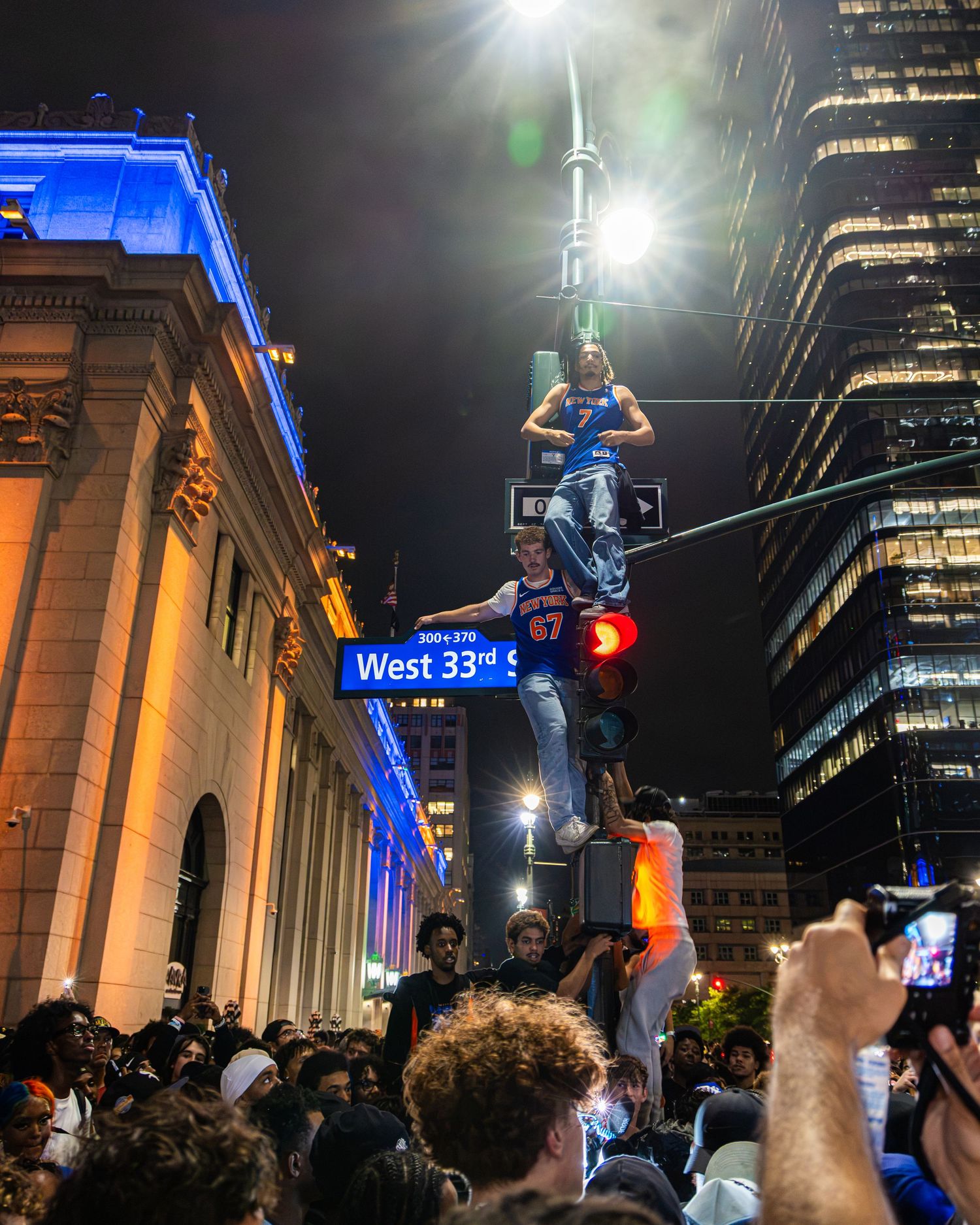 Crowd celebrating at night on West 33rd Street NYC, with people climbing a street sign post amid a large urban gathering.