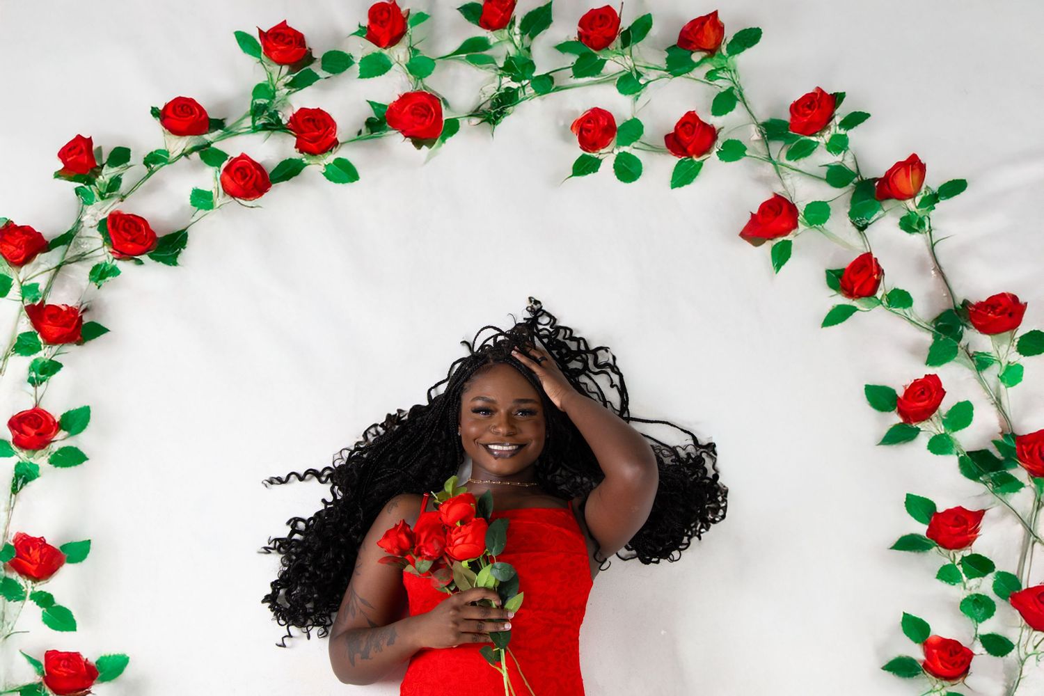 Woman in red dress lying among red roses on white background, holding bouquet, surrounded by floral arch.