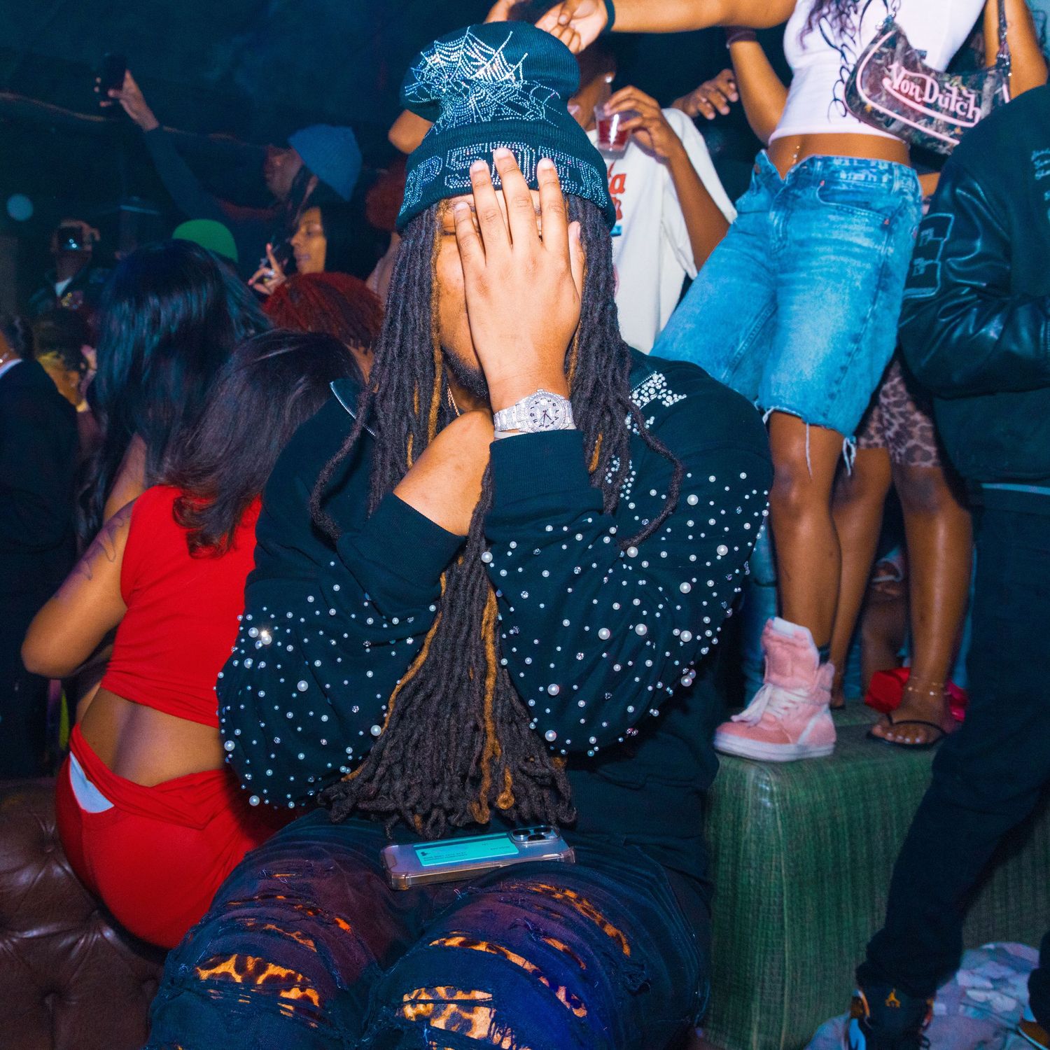 Person with dreadlocks in a sparkly jacket at a nightclub party with dancers in colorful outfits.