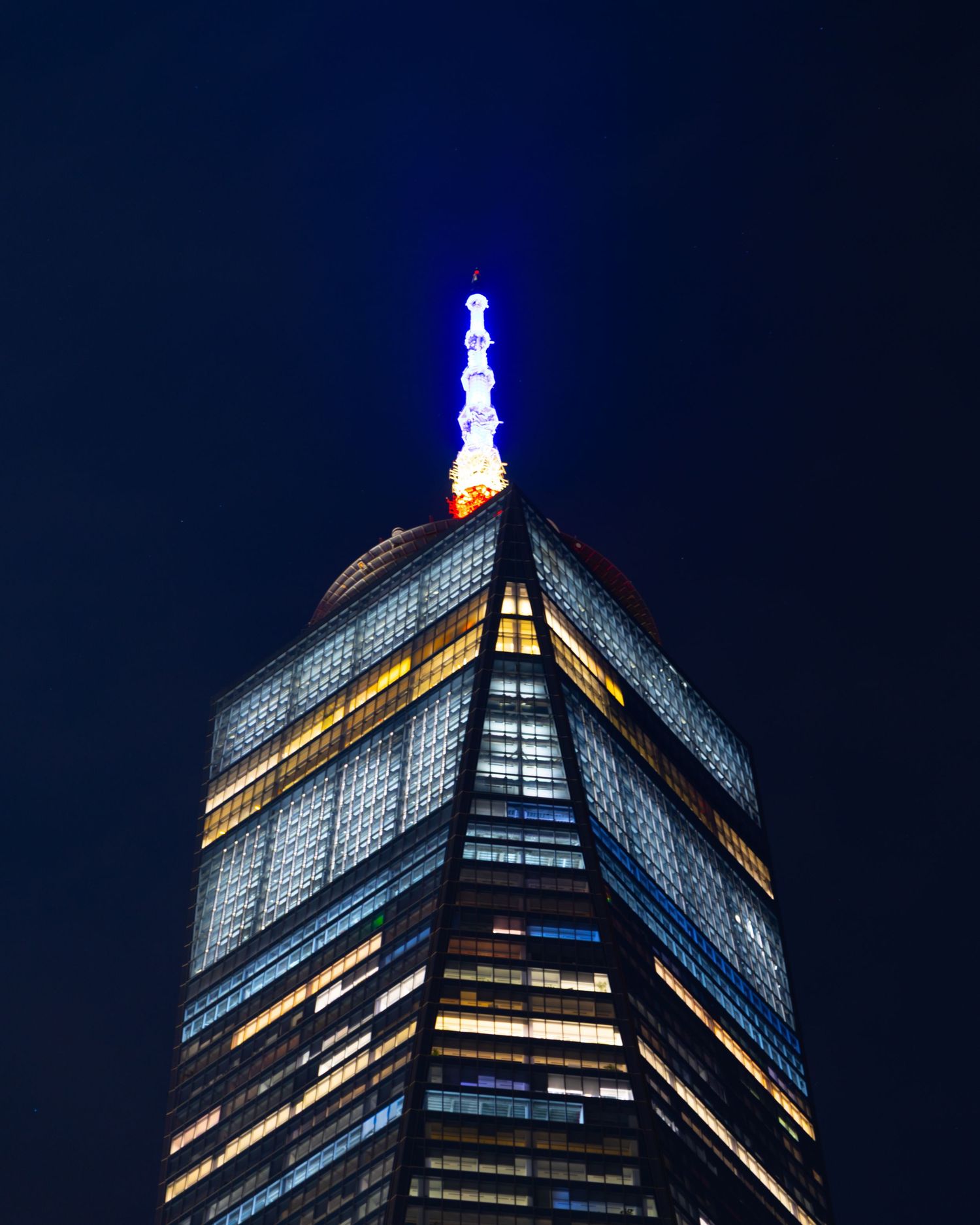 One World Trade Center illuminated at night with a glowing blue and white spire against a dark blue sky.