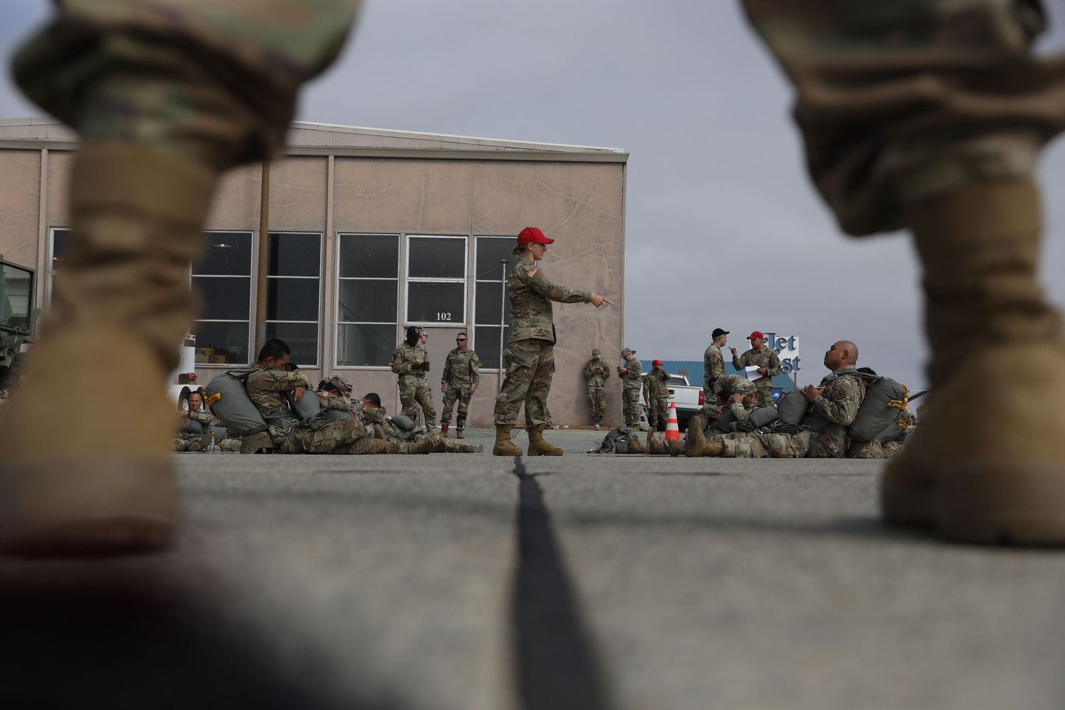 Military instructor in red cap briefing a group of soldiers sitting on pavement outside a building.