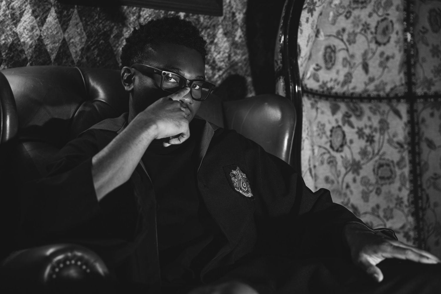 Black and white photo of a stylish man in sunglasses and blazer sitting in a leather chair.