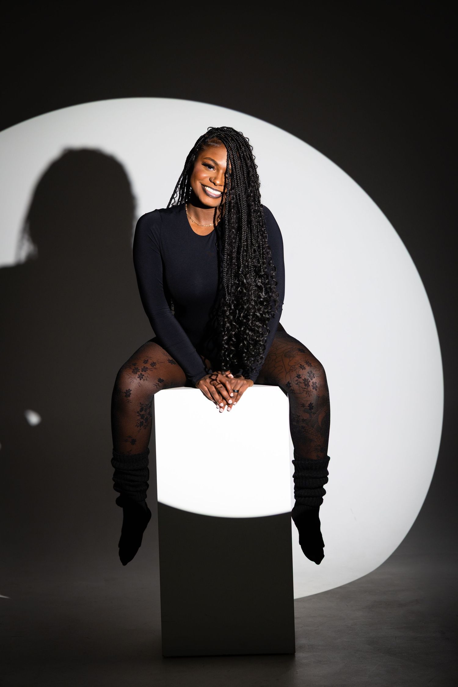 A smiling woman with long braids sits on a glowing white cube, wearing a black bodysuit and patterned tights.