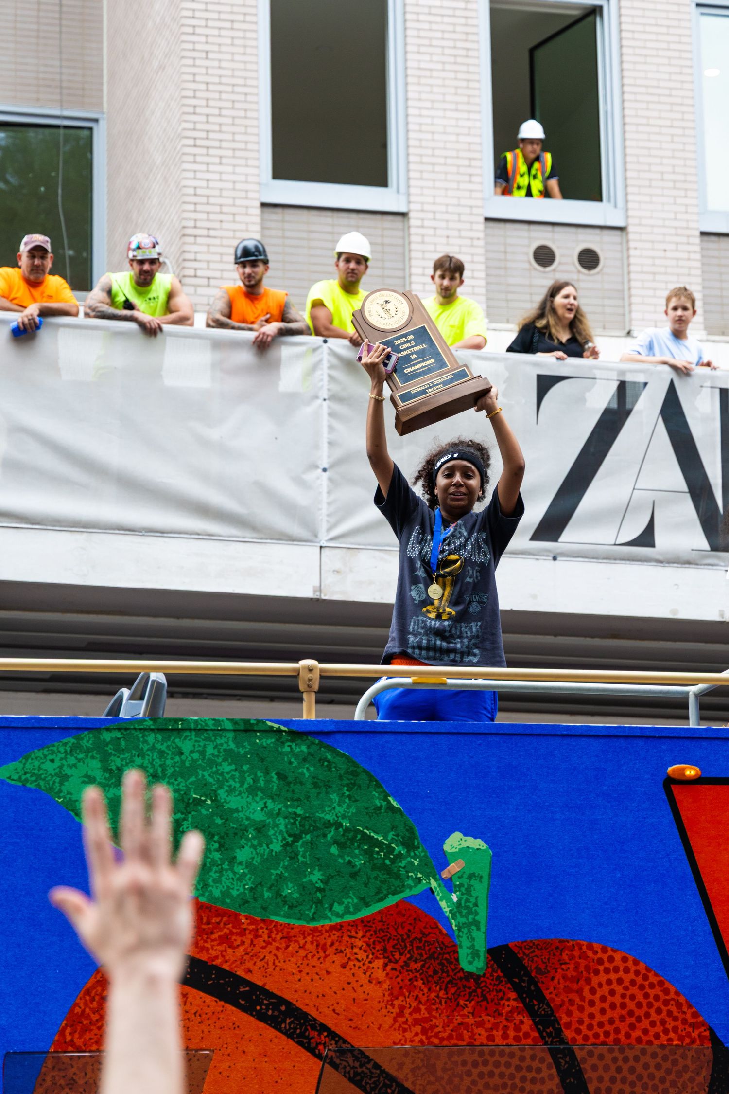 Athlete raises championship trophy on float during parade, cheered by construction workers on balcony above.