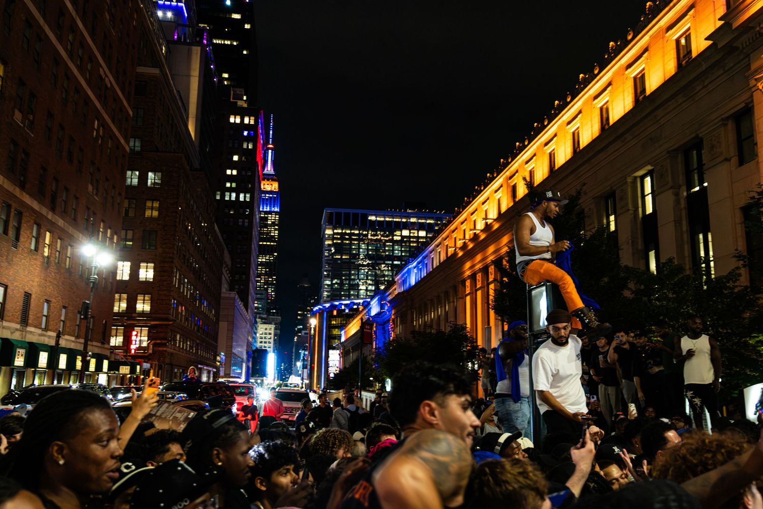 Nighttime street celebration with large crowd in urban city, illuminated by colorful building lights and skyscrapers in background.