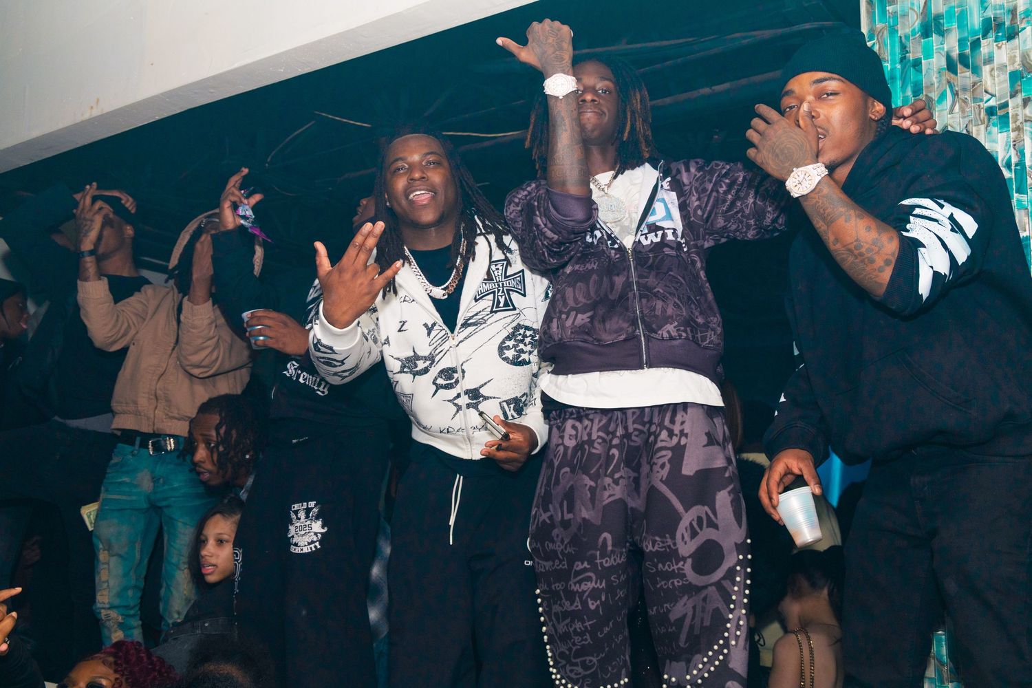 Four young men posing together at a nightclub party, wearing stylish streetwear outfits under blue lighting.