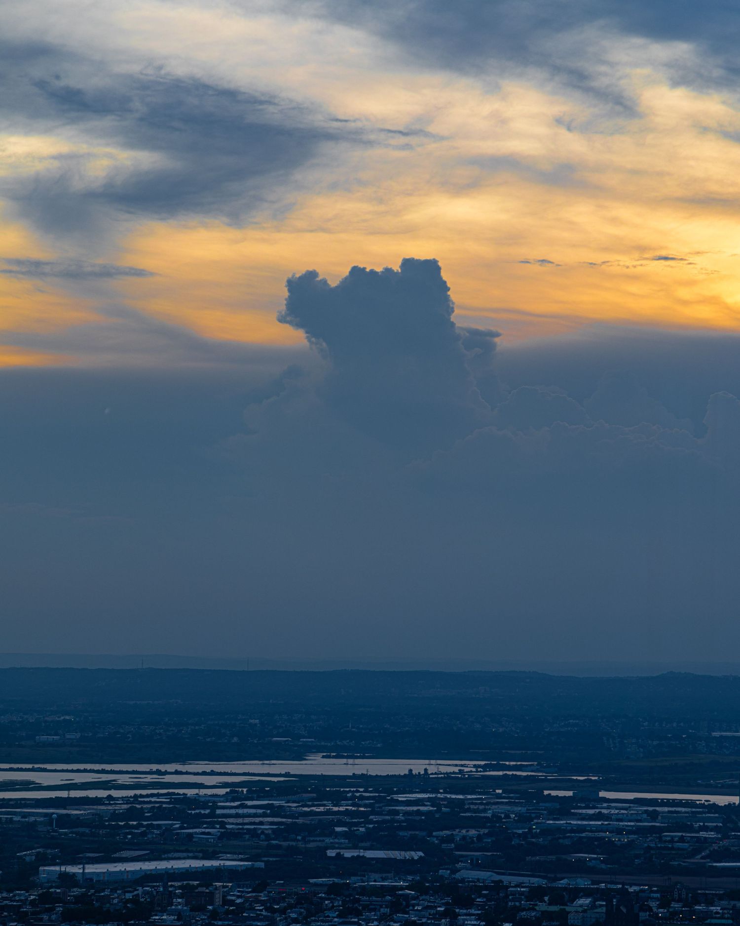 Dramatic sunset sky with a towering cumulonimbus cloud silhouetted against golden orange hues over a cityscape.