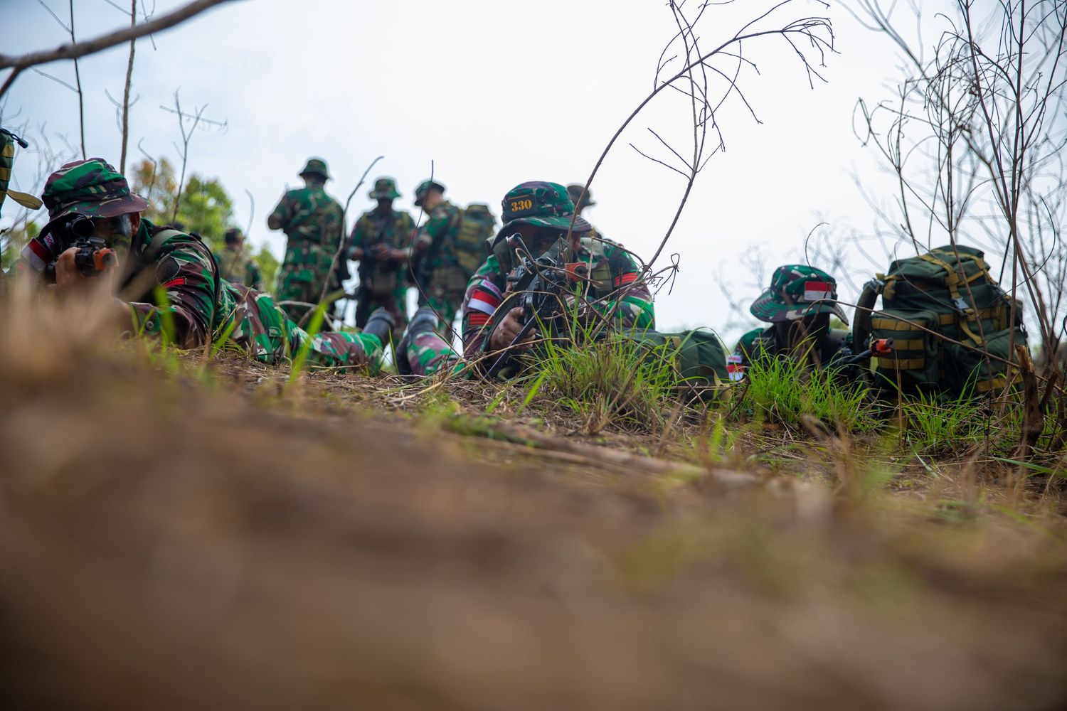 A group of soldiers in camouflage gear rest on a hillside during an outdoor military field exercise.