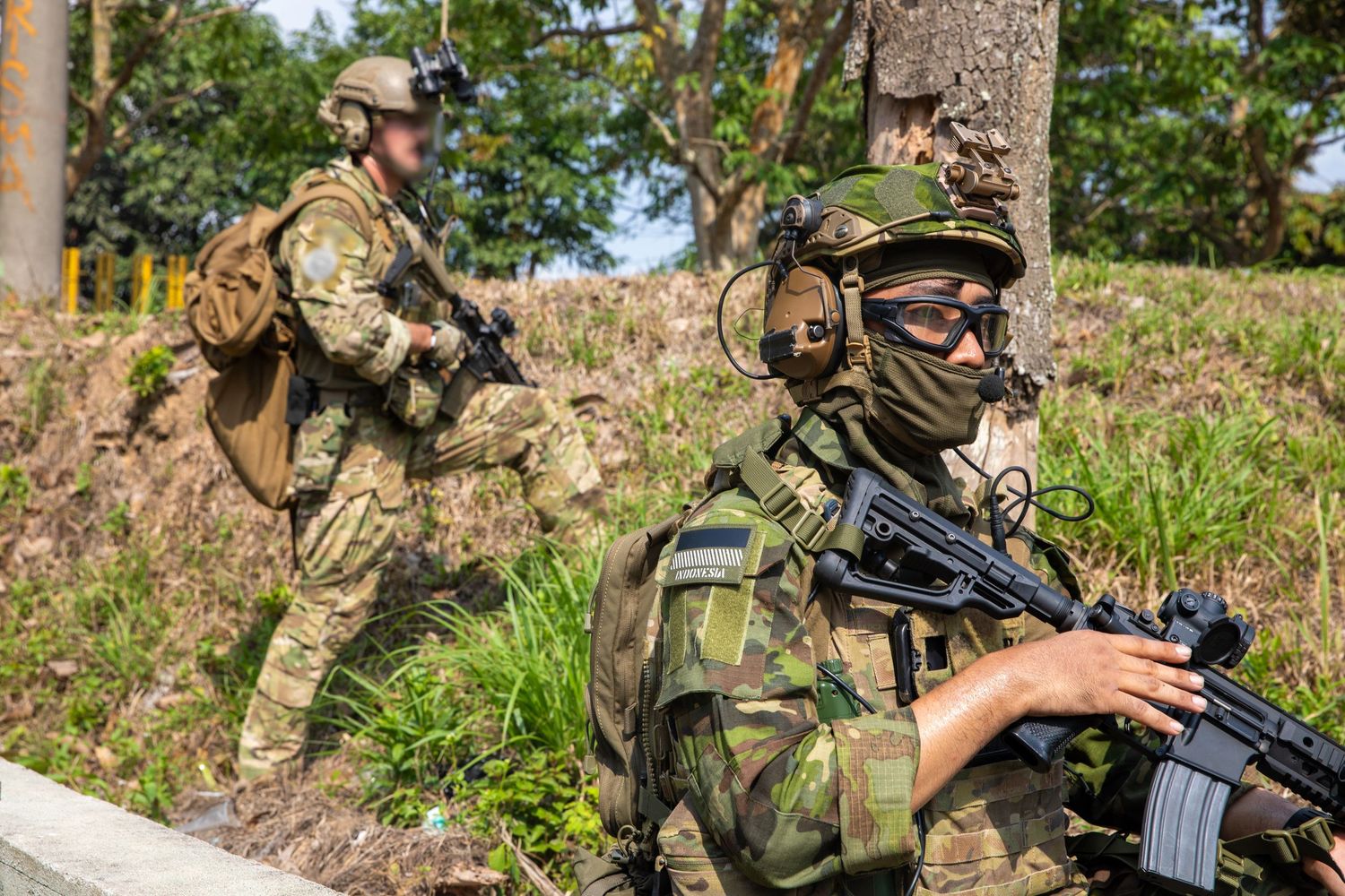 Two armed soldiers in camouflage gear patrol a wooded hillside during a military exercise.
