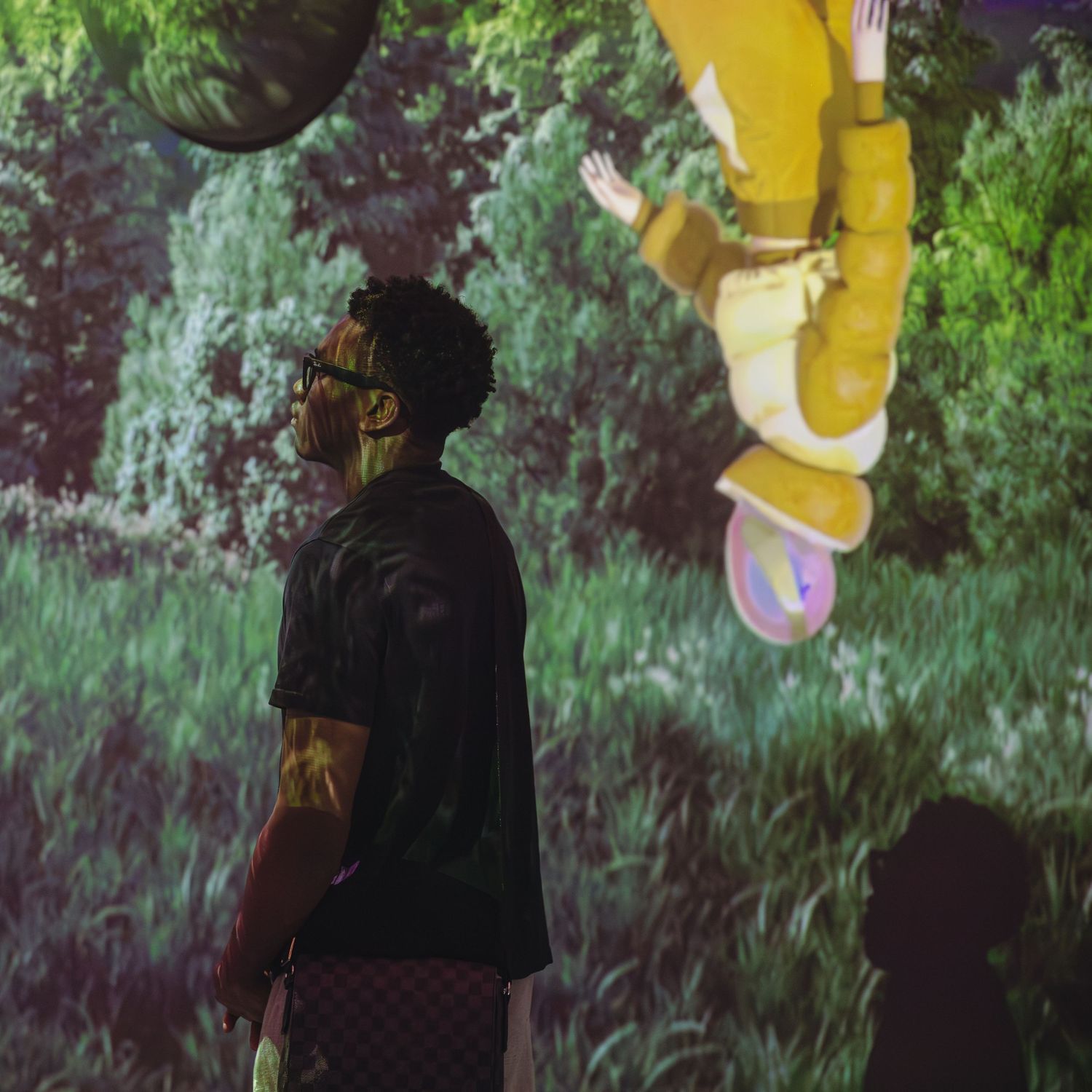 Man in black shirt views immersive digital art projection of cartoon figure in lush green forest setting.