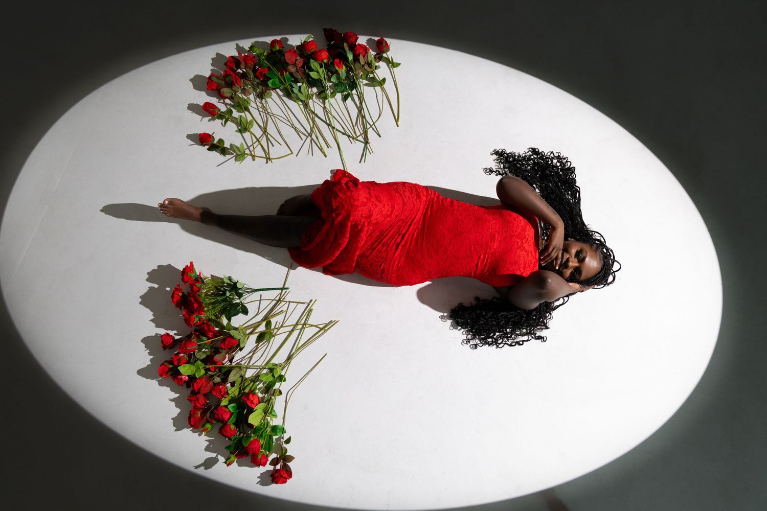 Woman in red dress lying on white oval surface surrounded by red roses, viewed from above.