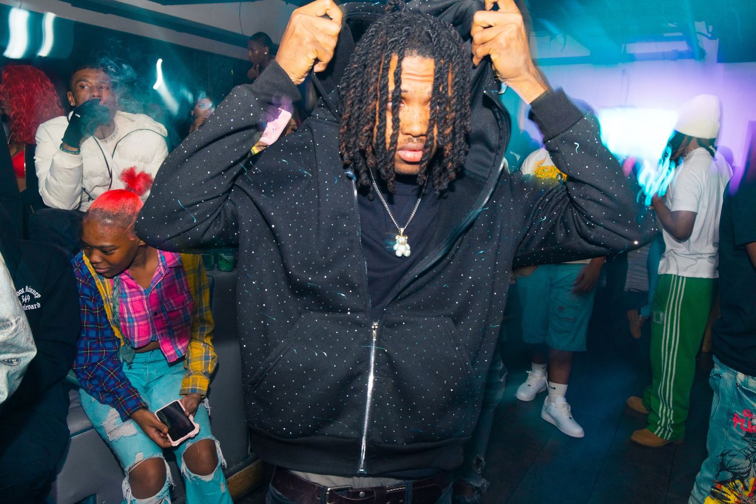Young man in sparkly black hoodie dancing at a vibrant nightclub party with colorful guests in background.