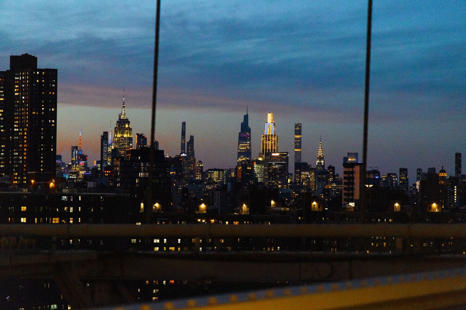 NYC skyline at dusk with illuminated skyscrapers including Empire State Building viewed from a bridge.