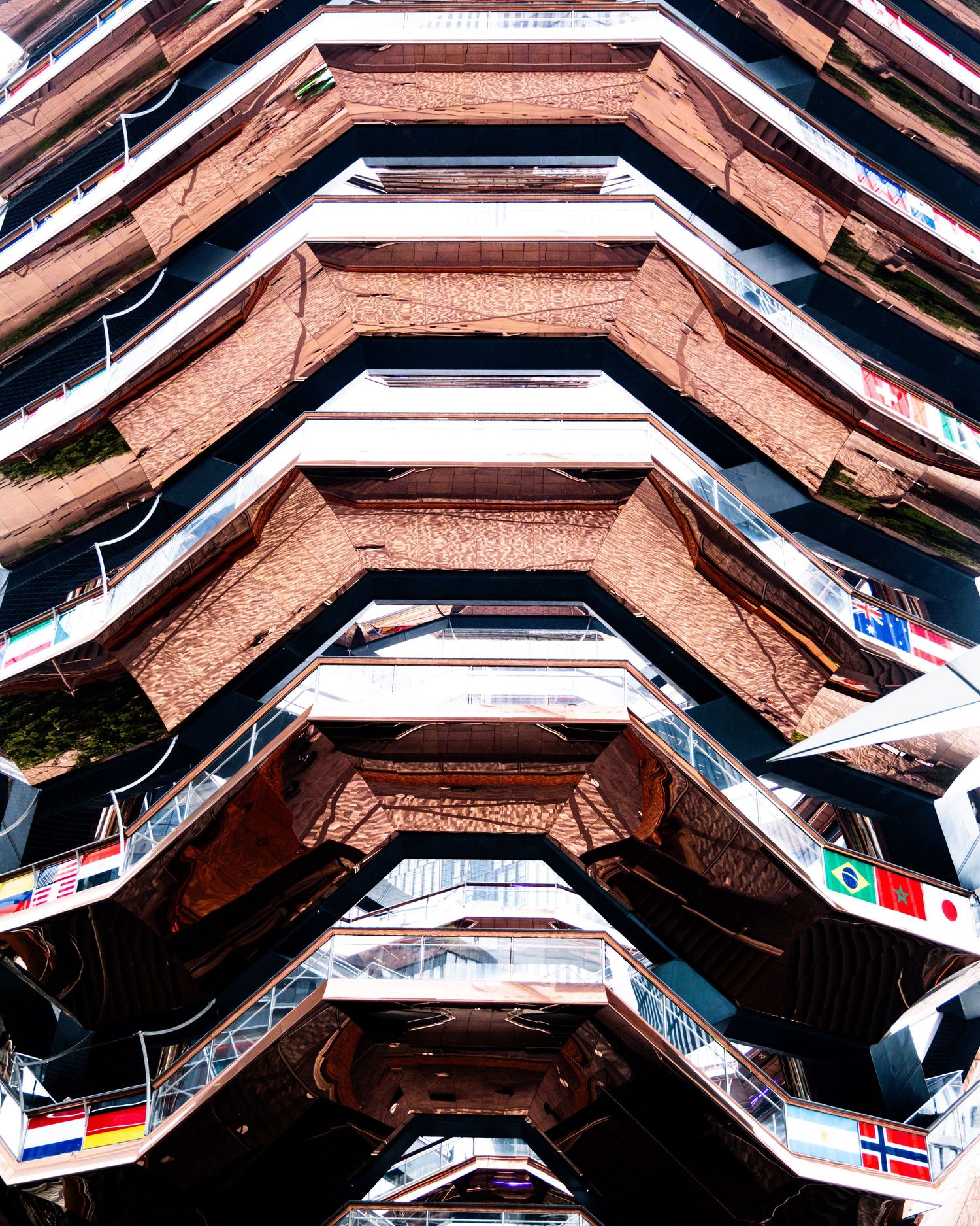 Looking up at The Vessel structure in Hudson Yards NYC, showing geometric honeycomb staircases with international flags.