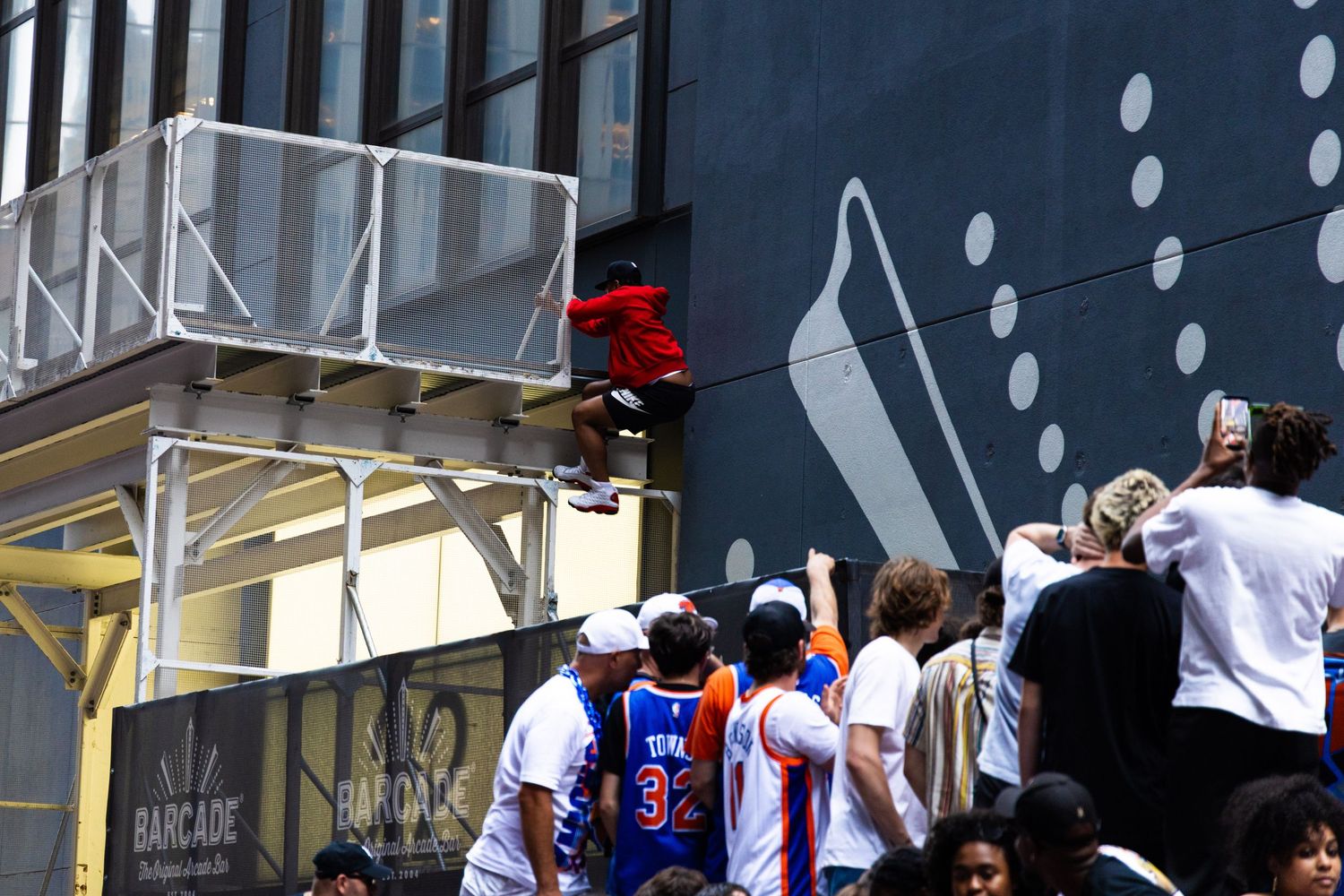 Person climbing urban building structure above crowd at outdoor street event with large mural backdrop.