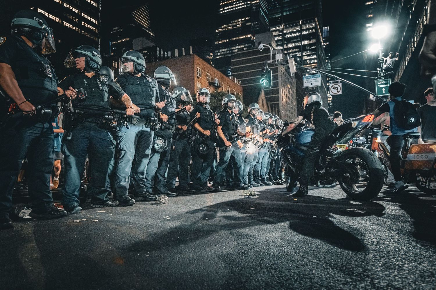 Police officers in riot gear face a motorcycle rider on a city street at night during a tense urban confrontation.