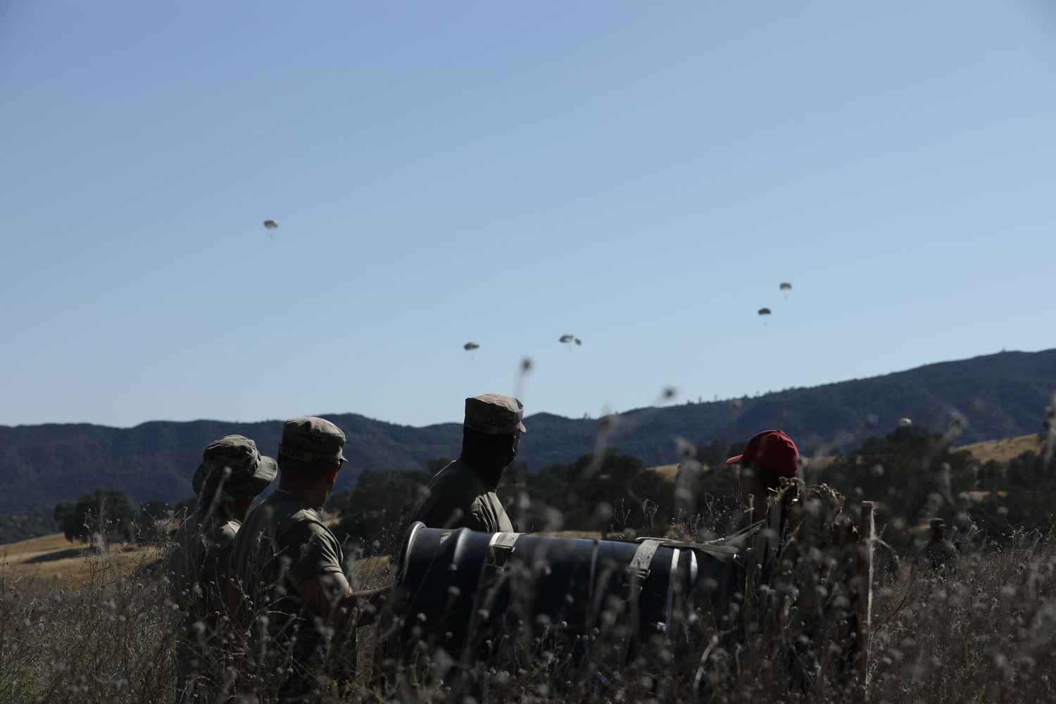 Military soldiers standing in field watching parachutes descend from aircraft over mountainous terrain.