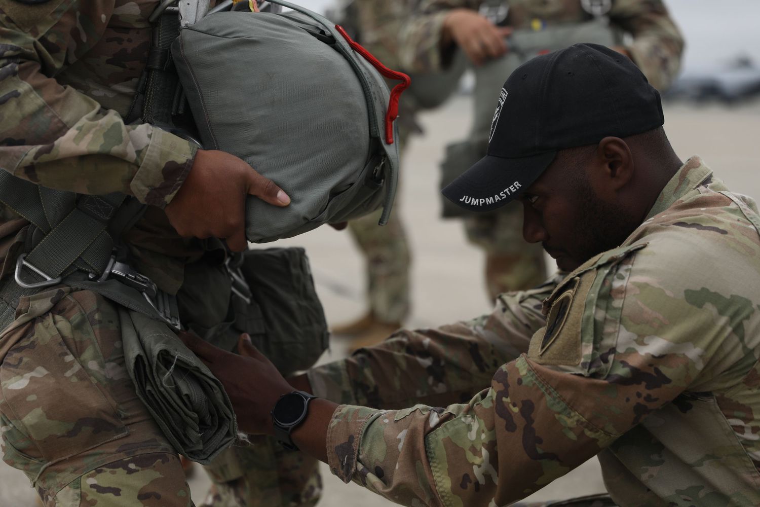 Military soldier checking and adjusting parachute gear of fellow soldier on airfield tarmac.