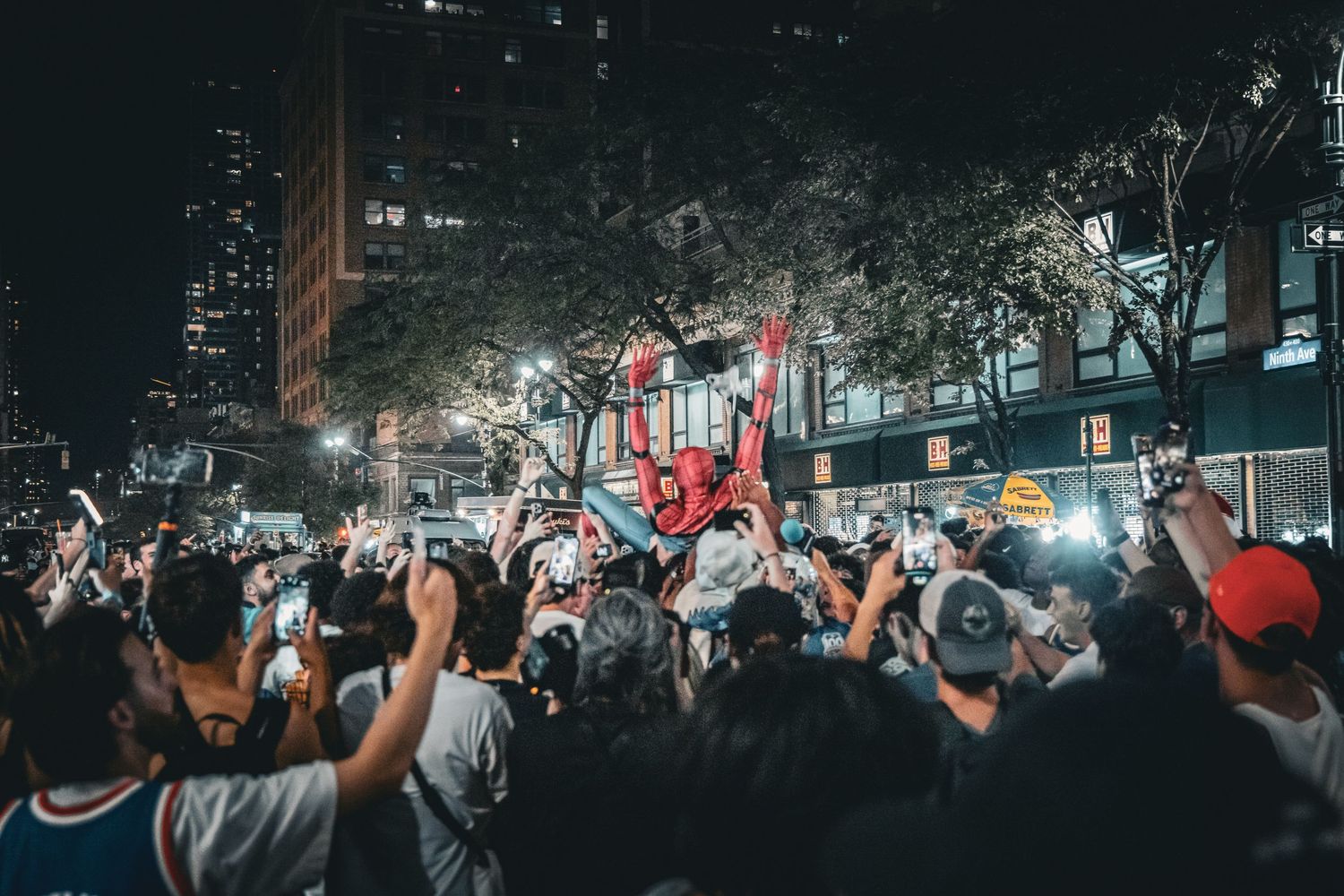 Large crowd celebrating at night on a city street with people raised on a structure amid bright lights.