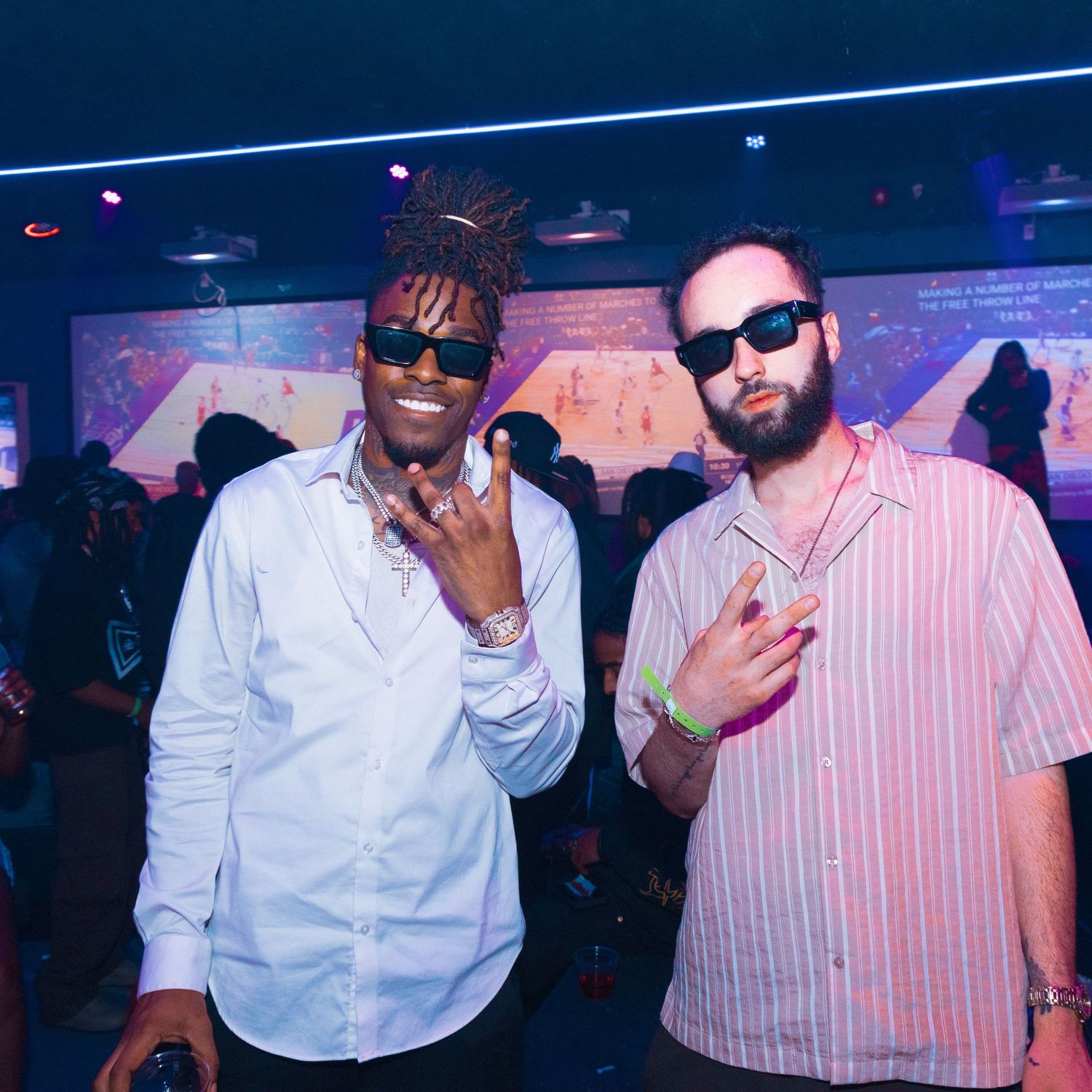 Two men posing together at a nightclub, one in a white shirt and one in a pink striped shirt, both wearing sunglasses.