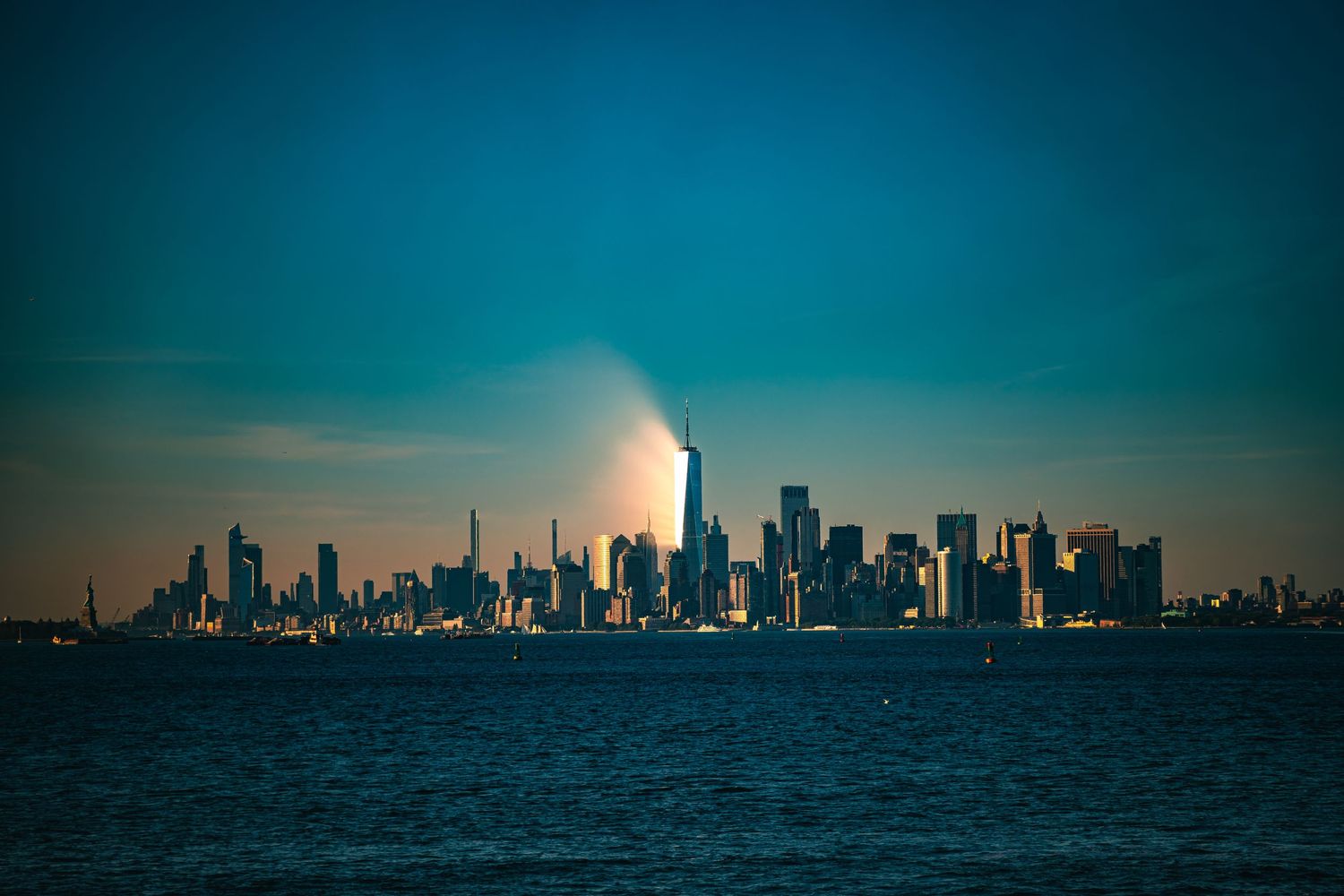 NYC skyline at dusk viewed from water, One World Trade Center illuminated against a teal sky.