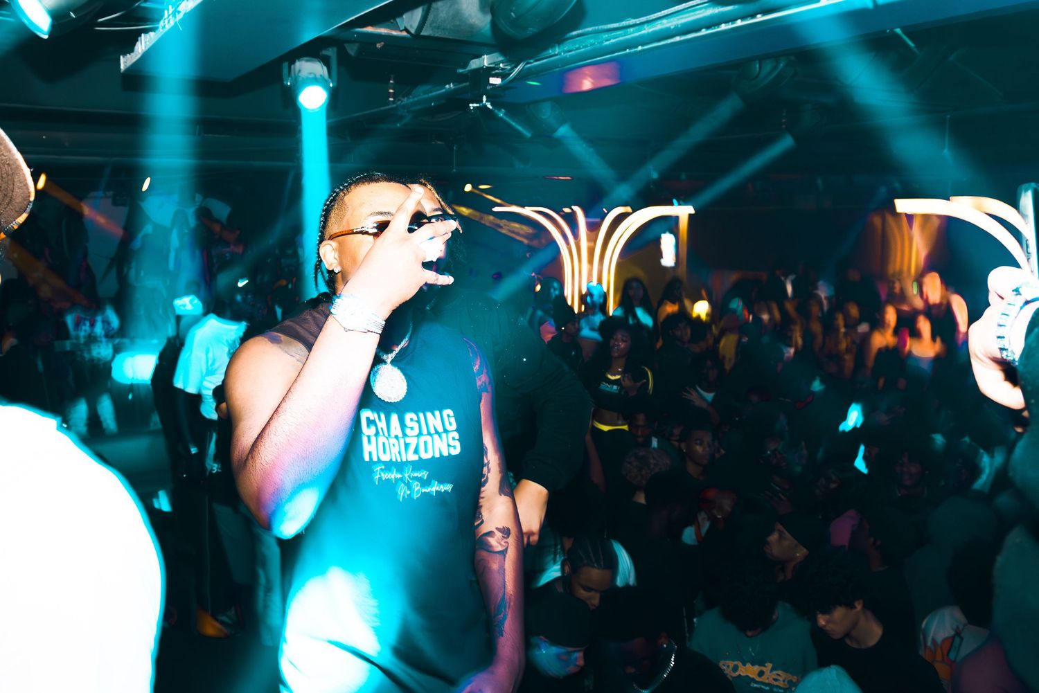 Performer on stage at a nightclub holding a microphone, wearing a black t-shirt, with crowd in background.