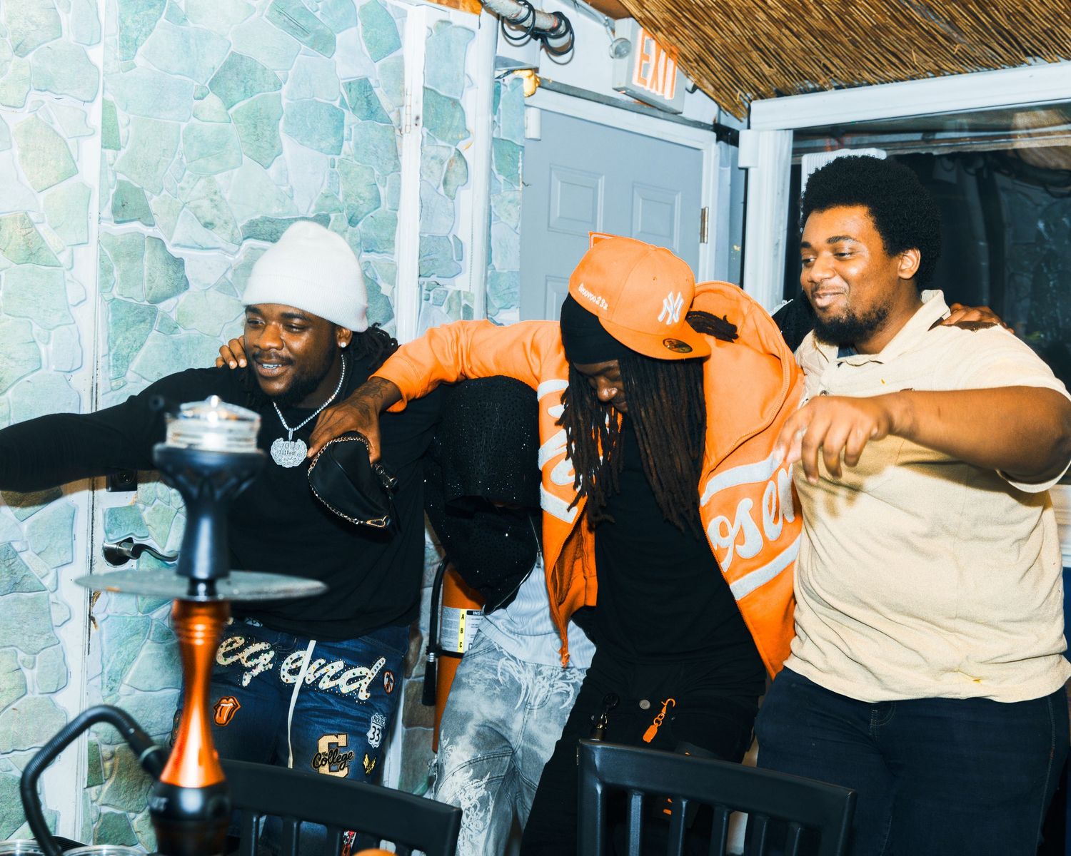 Three friends laughing and posing together indoors, one wearing an orange jacket and cap at a casual gathering.