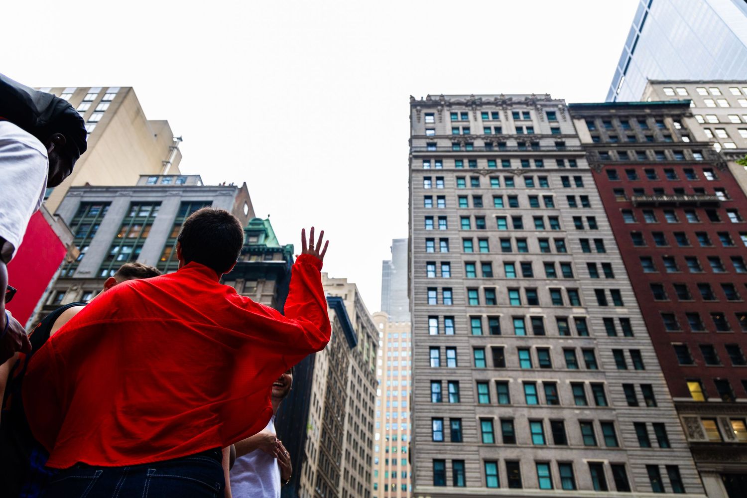 Person in red jacket gestures toward towering skyscrapers on a busy urban street in New York City.