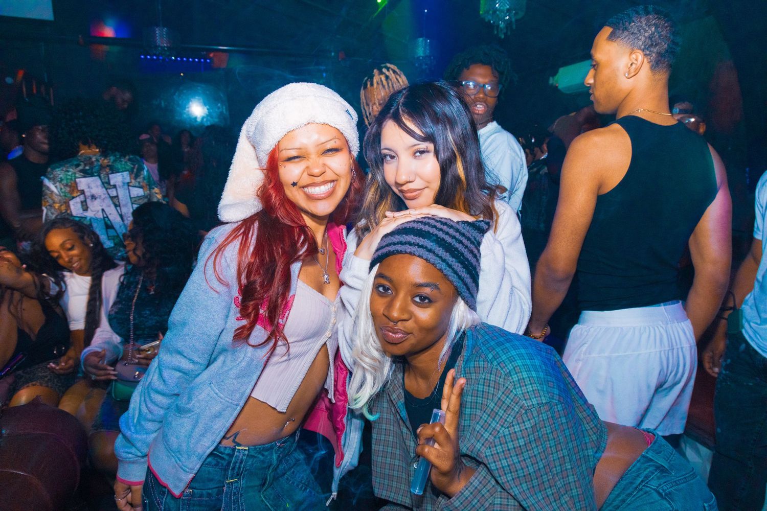 Three young women pose together smiling at a nightclub party with colorful lights and dancers in the background.