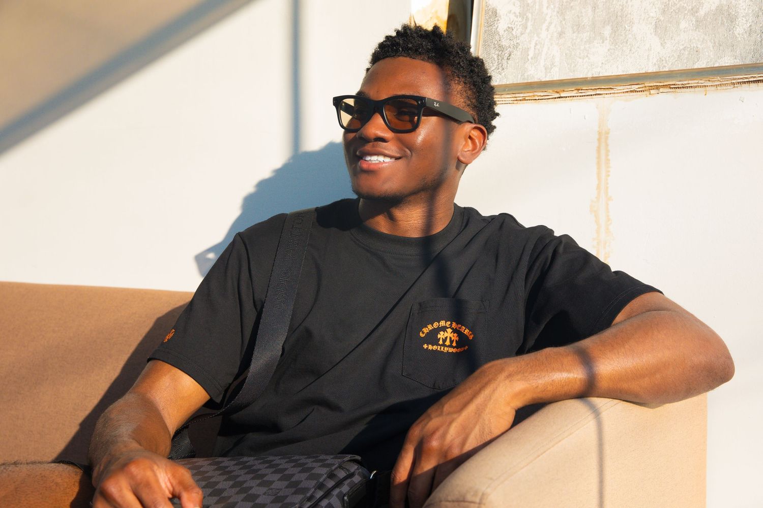 Young man smiling in black graphic tee and sunglasses, relaxing outdoors in warm sunlight.