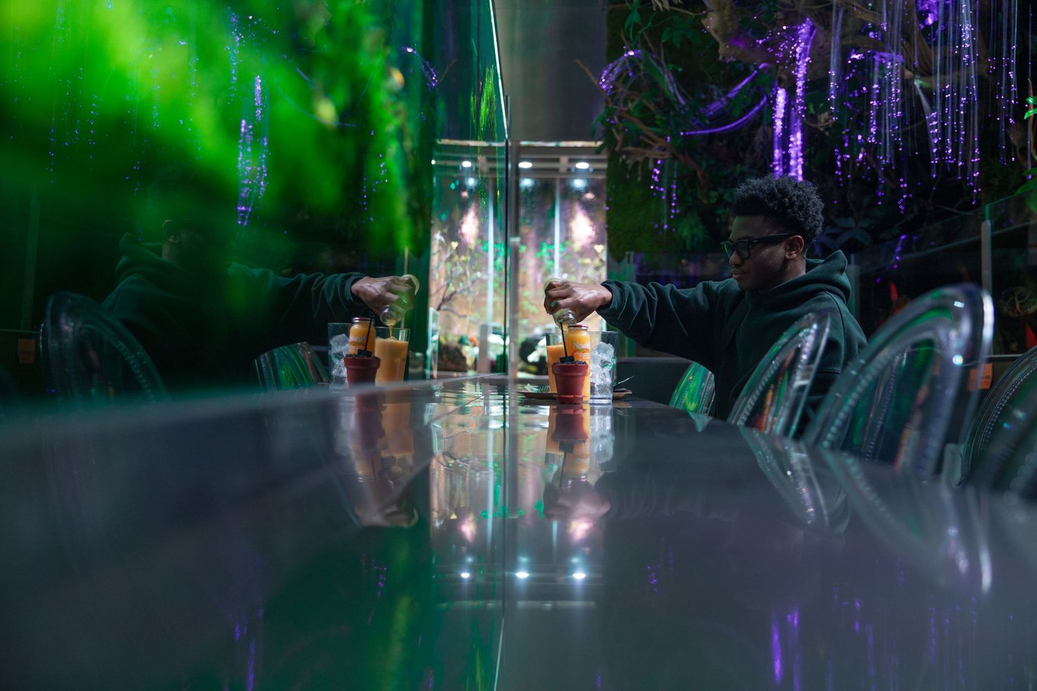 A man sits at a glowing green illuminated glass table in a neon-lit bar with colorful decorations.