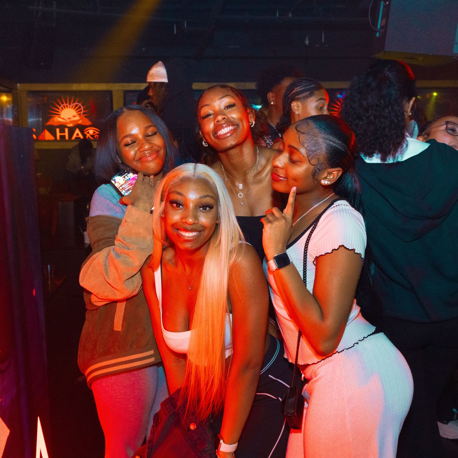 Group of young women posing and smiling together at a nightclub with warm orange lighting.