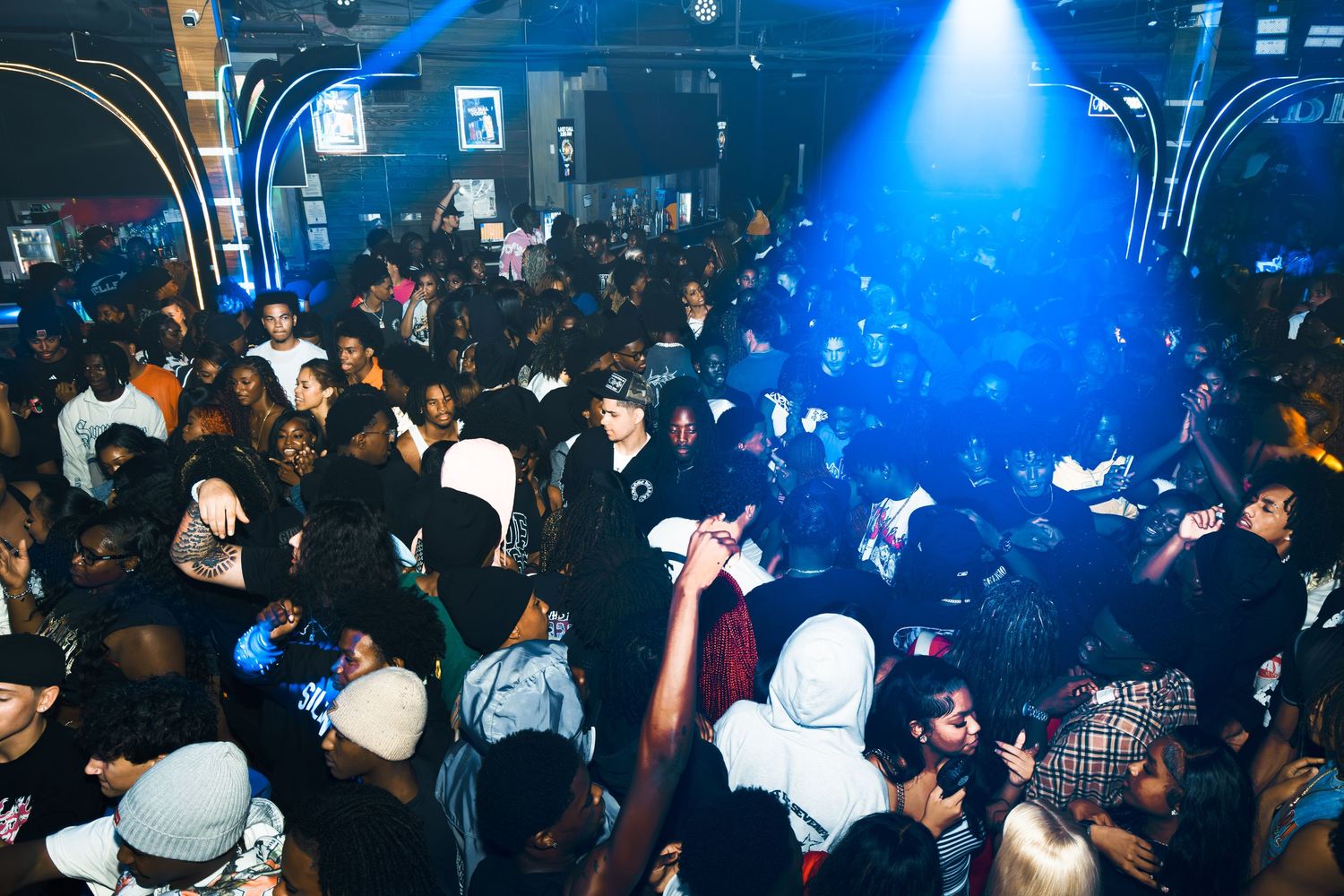 Crowded nightclub party with blue stage lighting, diverse group of people dancing and socializing on the dance floor.