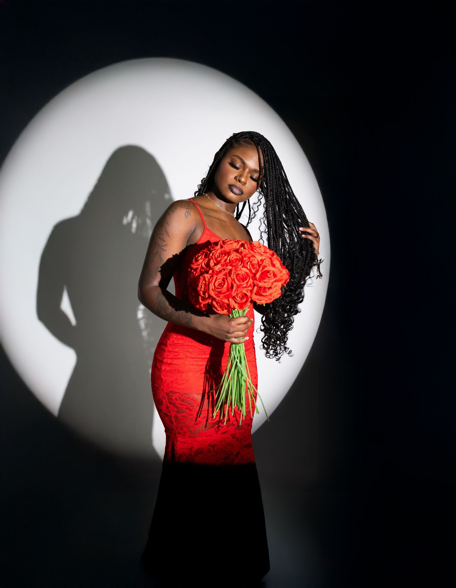 Woman in red gown holding red roses bouquet, posed against dramatic circular spotlight with shadow on dark background.