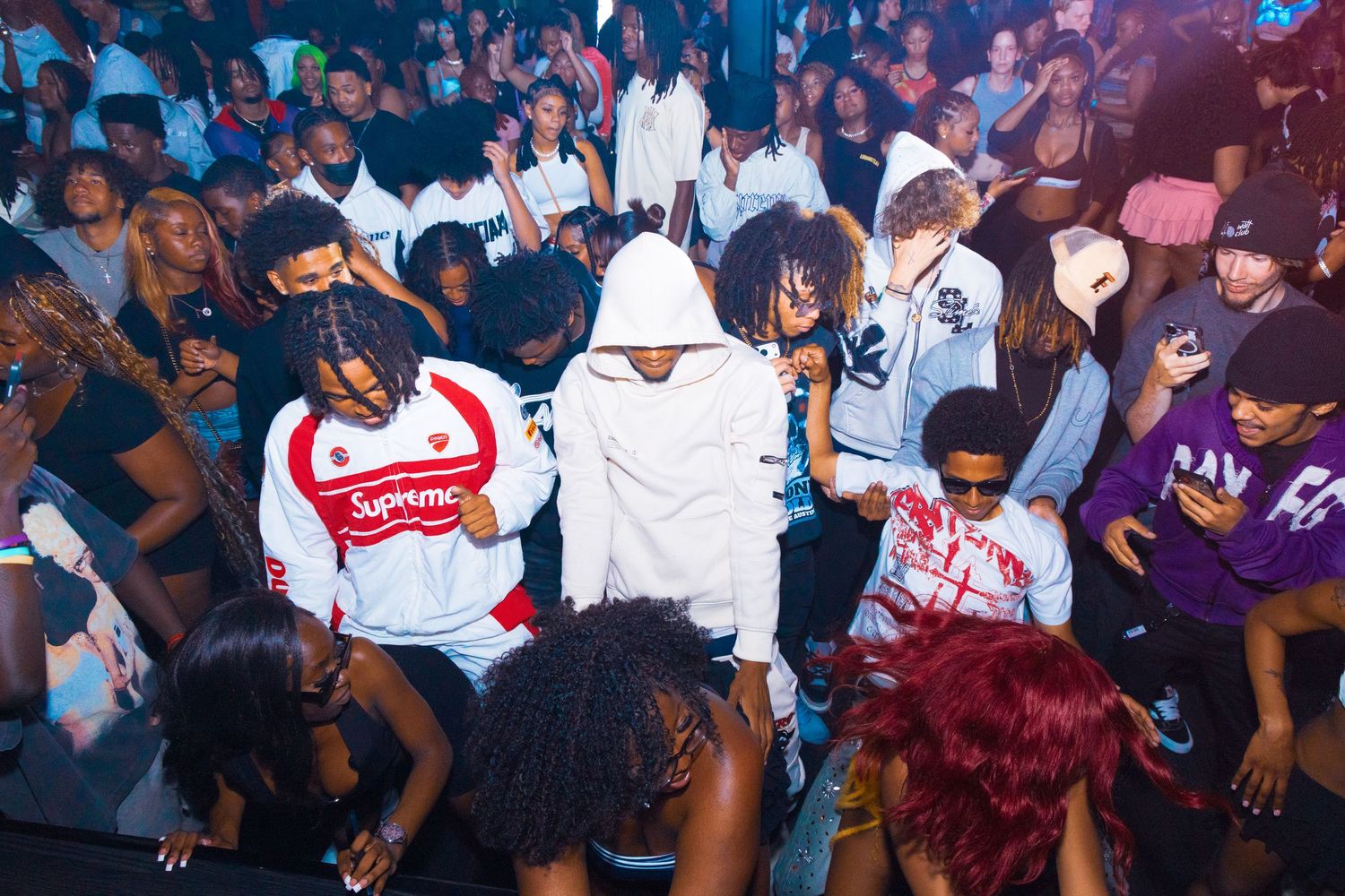 Young people dancing and socializing at a crowded nightclub party, wearing streetwear including Supreme shirts.