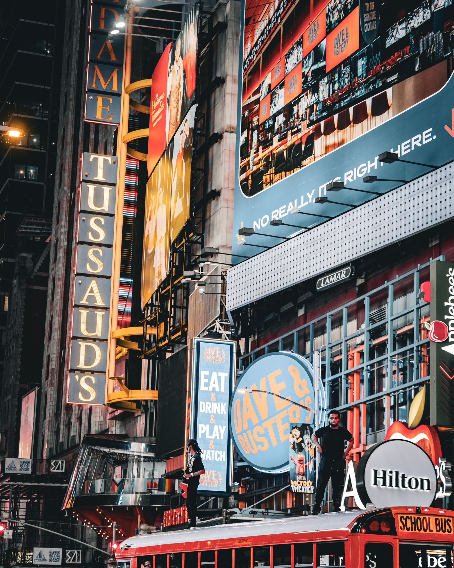 Vibrant Times Square NYC night scene featuring Madame Tussauds sign, Hilton hotel bus, and colorful illuminated billboards.