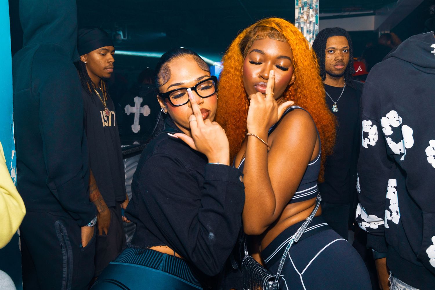 Two stylish women posing together at a nightclub event, one wearing glasses and a black hoodie, the other with orange curly hair.