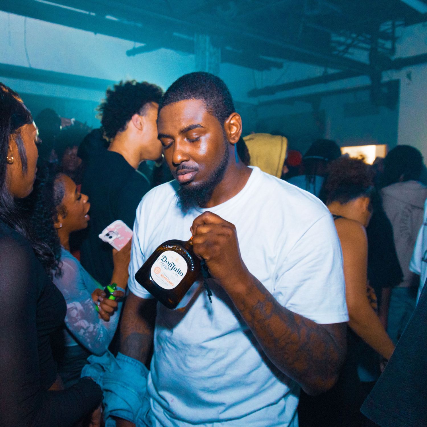 Man in white t-shirt holding a bottle at a crowded nightclub party with blue lighting.