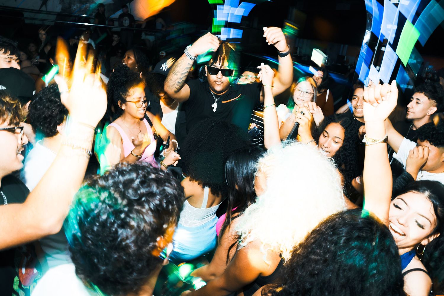 Energetic nightclub party scene with DJ performing on stage surrounded by excited dancing crowd under colorful lights.