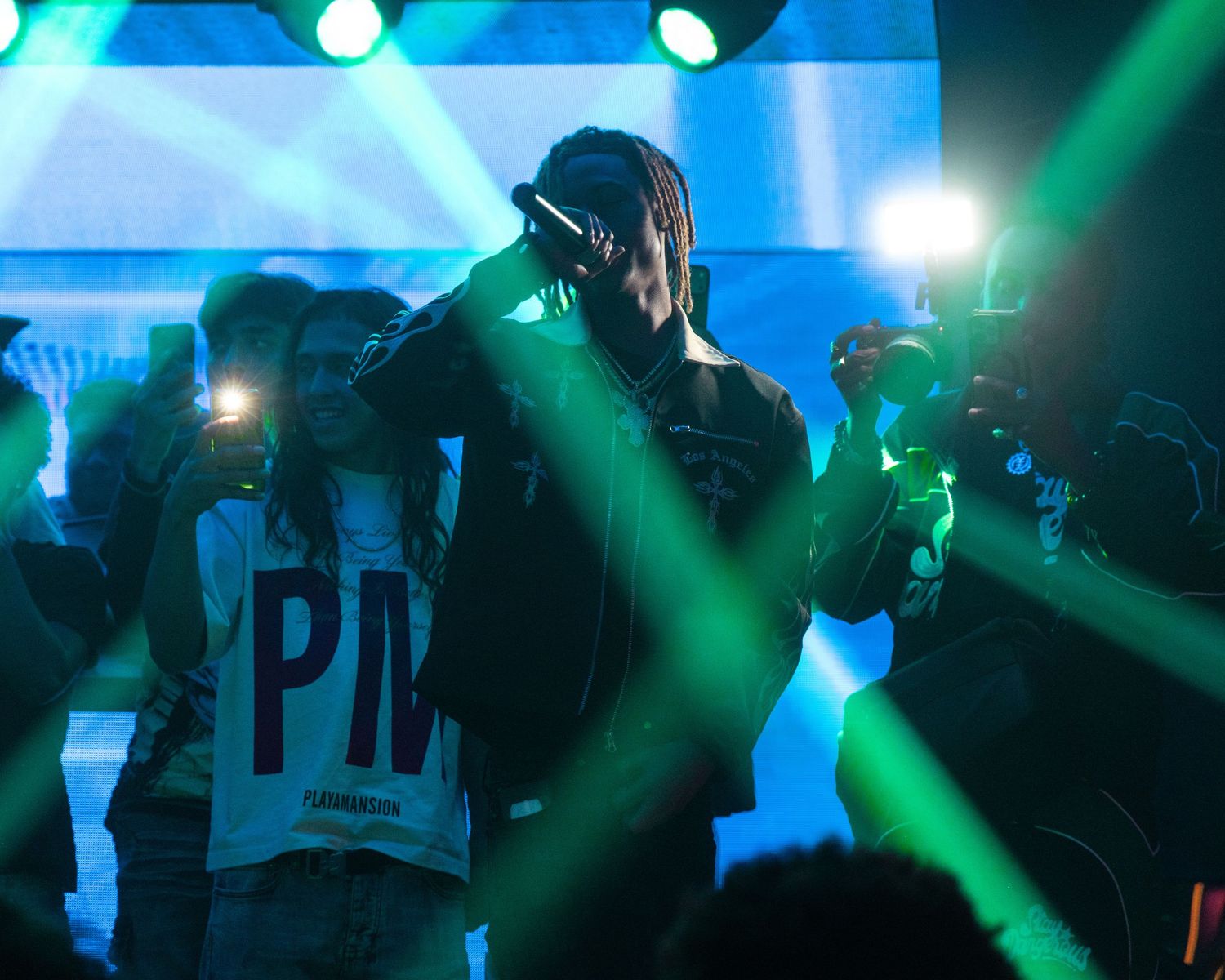 Rapper performing on stage with green laser lights and crowd holding phones at a concert.