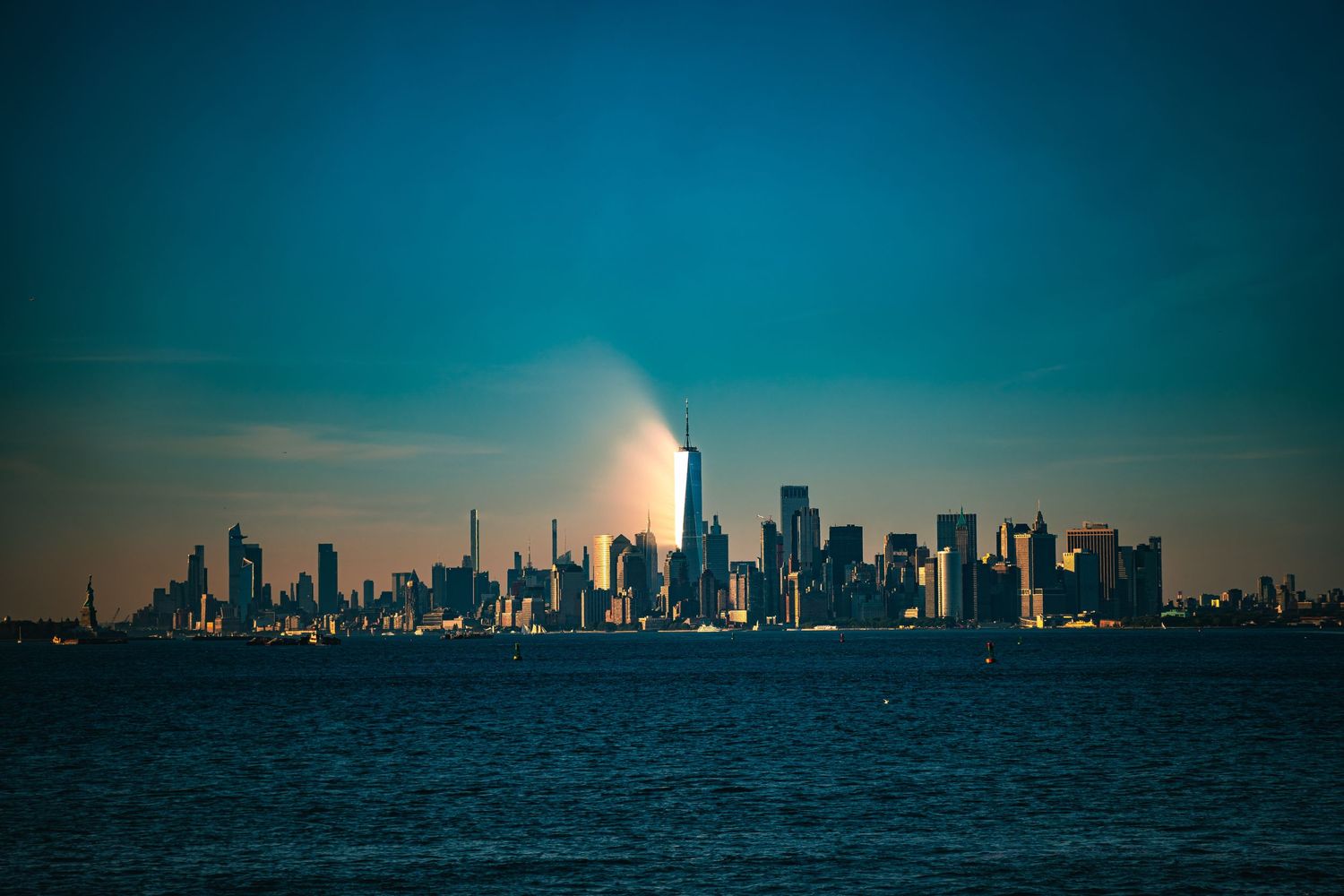 New York City skyline at dusk viewed from the water, with One World Trade Center illuminated against a teal sky.