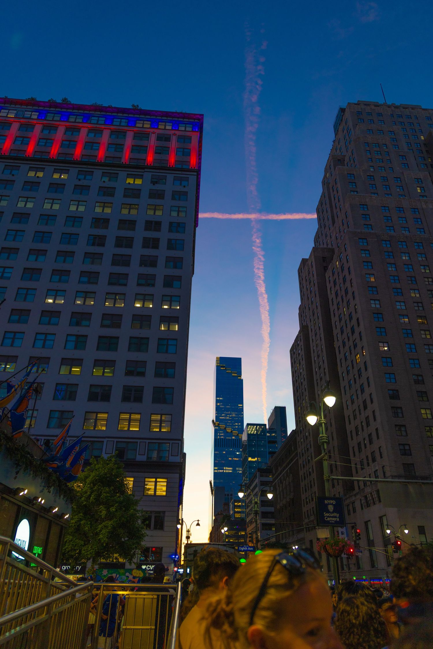NYC skyline at dusk with contrails forming a cross between illuminated skyscrapers and a crowd below.