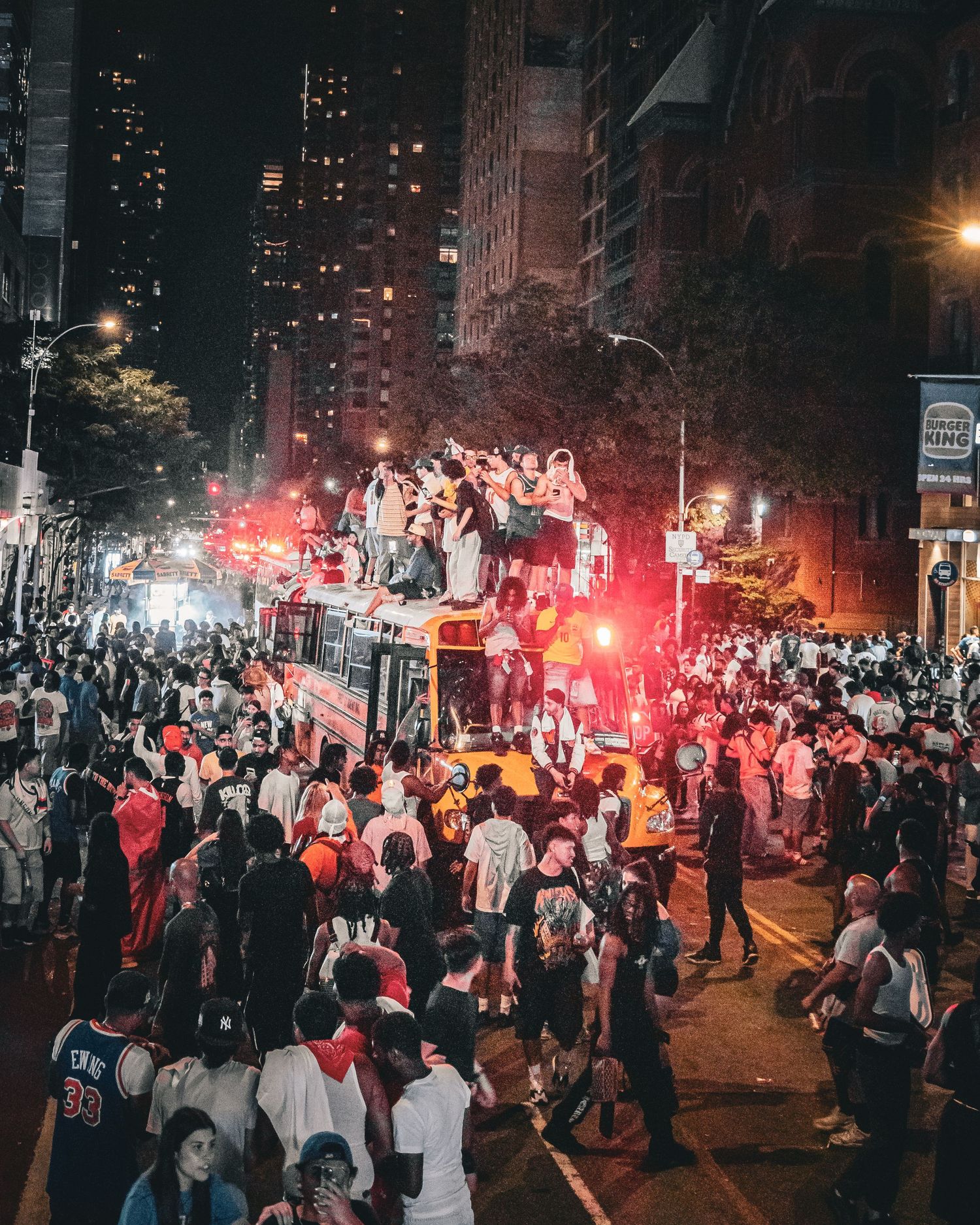 Massive nighttime street celebration with crowds surrounding a double-decker bus, illuminated by red lights in an urban city setting.