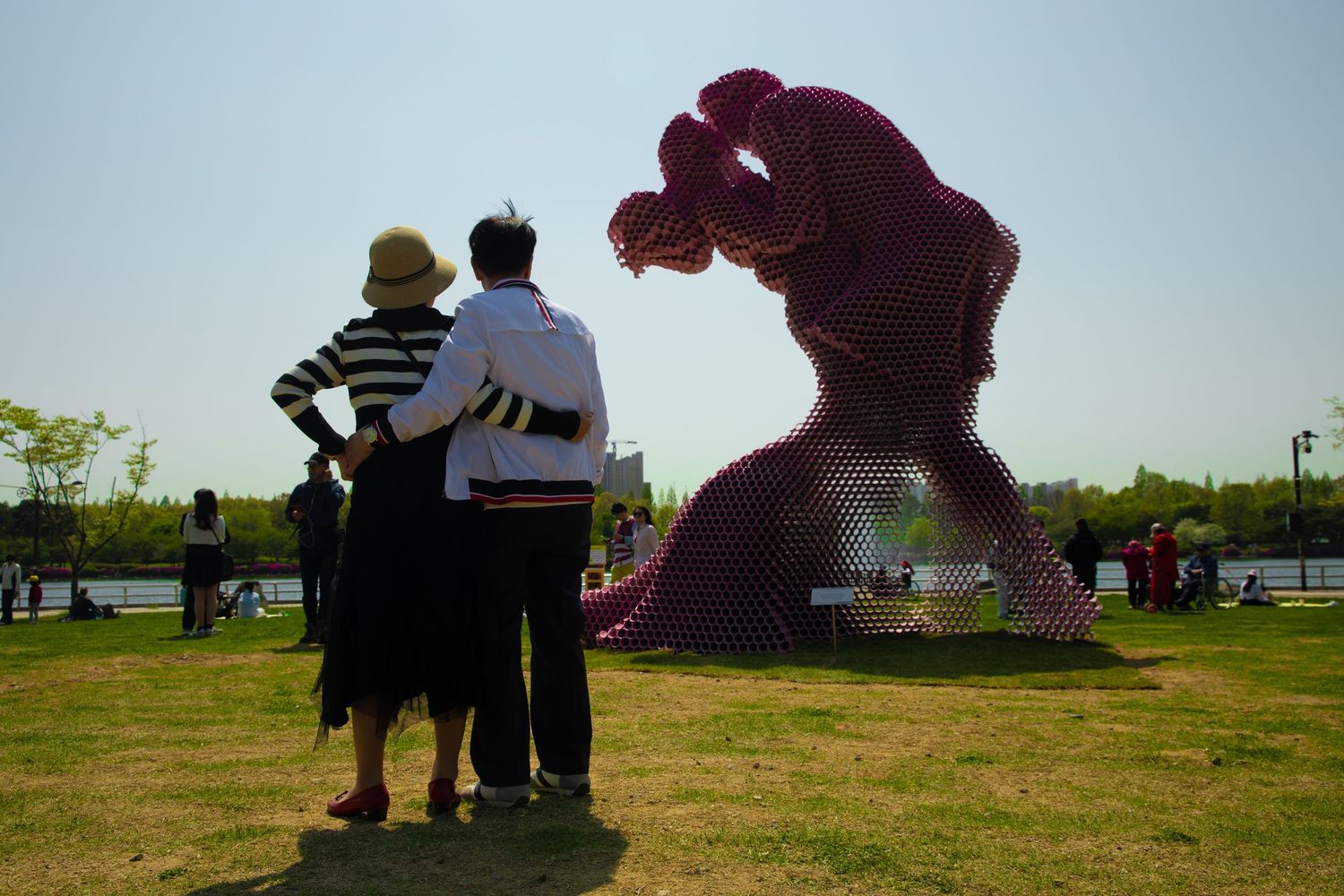 Two people stand viewing a large red floral sculpture in a park on a sunny day.