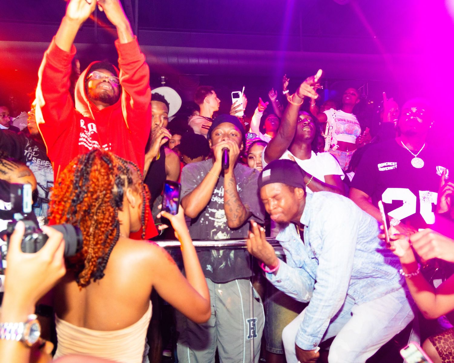 Energetic nightclub party scene with crowd dancing under red lights, people raising hands and celebrating.